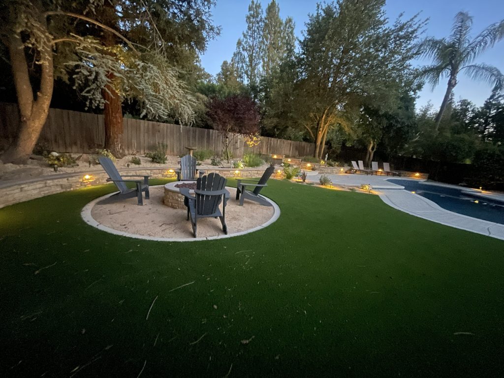 Custom fire pit gathering area surrounded by decorative retaining walls, landscape lighting, and outdoor living spaces in the completed Wedemeyer backyard renovation by Sunset Construction.